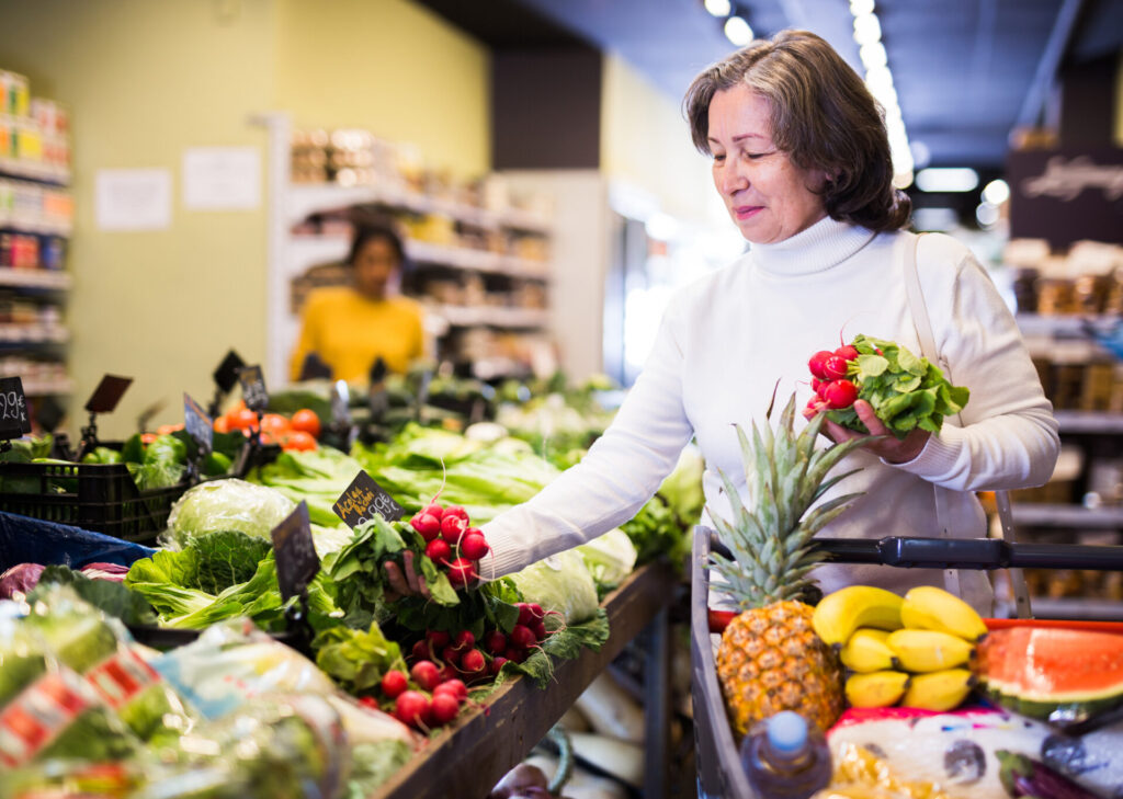 Older woman grocery shopping in the produce aisle. She is evaluating her radish options