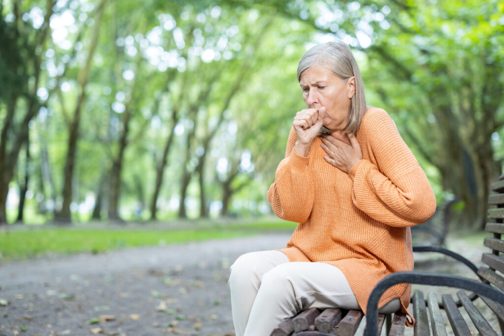 Woman sitting on a bench in the park. She is pressing on her chest with one and and coughing into her fist on her other hand