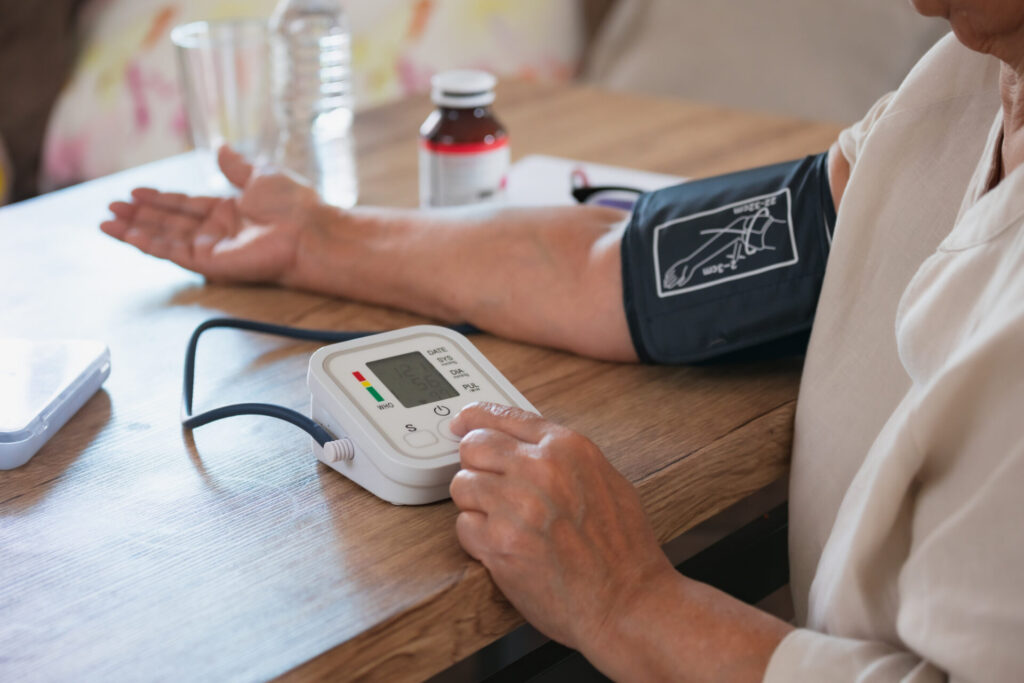 Older woman at her kitchen table with a blood pressure cuff wrapped around her arm. She is checking her blood pressure levels.